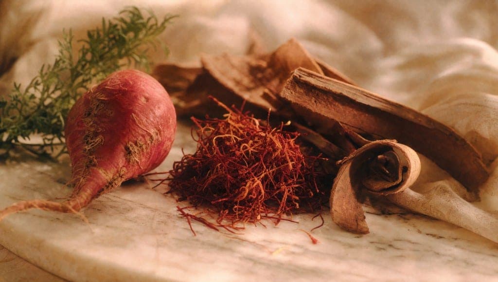 Une racine de maca rouge à fanes vertes, un tas de filaments rouges et des plaques d'écorce dont une enroulée, sur un marbre clair.