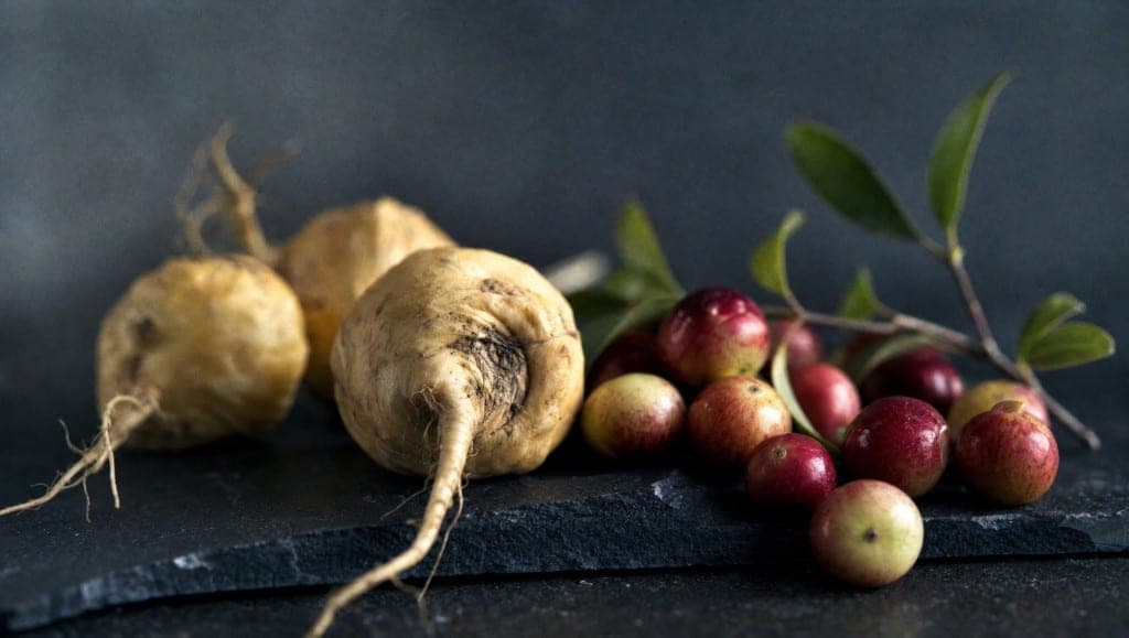 Trois racines de maca beiges sur une ardoise sombre, des baies de camu camu rouges et vertes et un rameau feuillu à côté.
